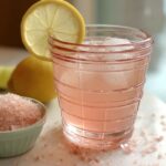 Refreshing pink salt weight loss drink served in a clear glass with ice cubes and a lemon slice, next to a bowl of Himalayan salt